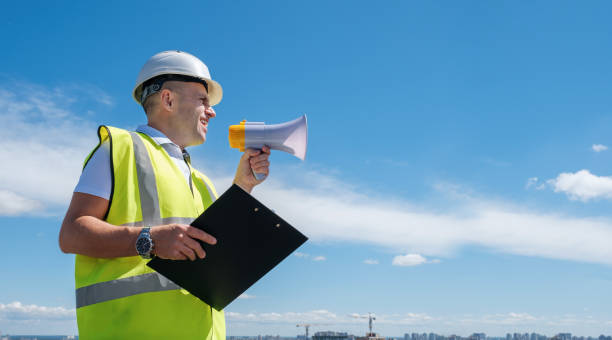 Construction safety communication - Builder, foreman or architect in a white helmet and a reflective vest shouts into a megaphone 